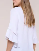 Blusa blanca de crepe crinkle con escote en V, media abotonadura frontal con botones nacarados, alforzas verticales en la pechera y mangas tres cuartos acampanadas con detalle de encaje horizontal.