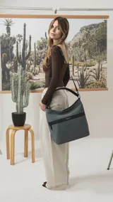 Cartera tipo hobo de tamaño grande en color beige, con detalles en negro. Presenta una franja horizontal negra con el logo de la marca, asa corta de hombro y correa larga desmontable. Incluye bolsillo frontal con cierre oculto y bolsillo trasero.