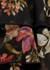 Blusa negra semitransparente con estampado floral en tonos rojos y rosas, volantes en el bajo, cuello redondo, manga larga abullonada y cierre de lazo en el cuello.