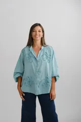 Blusa de color turquesa con cuello mao, cierre frontal de botones y mangas tres cuartos abullonadas. Presenta un diseño con bordados florales en relieve y detalles calados en el frente.