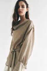 Camisa de cuello fluido y manga larga con puños anchos, de corte recto y largo medio, confeccionada en tejido rústico en tono beige con sutil textura jaspeada. Cuenta con un cinturón ajustable del mismo género para entallar la cintura.