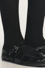 Zapato tipo bailarina de acabado acharolado en color negro, con punta redonda y suela plana. Presenta una tira ajustable con hebilla metálica y bandas elásticas cruzadas en el empeine.