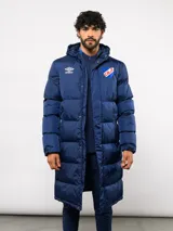 Parka larga acolchada color azul marino con capucha, cierre frontal con botones a presión y bolsillos laterales. Presenta el logo de Umbro y el escudo del Club Nacional de Football bordados en el pecho.