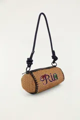 Bolso tipo bandolera de formato cilíndrico, confeccionado con material trenzado en color tostado, con detalle de bordado frontal que dice 'Rio' acompañado de una palmera y un tucán. Cuenta con una correa larga de cuerda oscura con nudos ajustables y cierre superior.