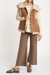 Chaleco estilo gamulán de corte oversize, confeccionado en material tipo gamuza color camel con interior y bordes de corderito sintético color crema. Presenta cierre frontal completo, bolsillos laterales y detalles de tiras decorativas en los hombros.