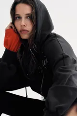 Sudadera negra con capucha, cierre frontal de cremallera metálica y mangas largas con puños. Presenta un diseño corto con bolsillo tipo canguro y detalle de texto 'California' aplicado con brillos en el frente.
