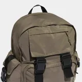 Mochila Adidas de diseño utilitario en color marrón, con compartimento principal amplio, bolsillo frontal con solapa y hebillas ajustables, y bolsillos laterales con solapa. Presenta el logo del trébol en la parte inferior frontal y detalles en negro.