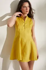 Vestido corto de lino color mostaza, sin mangas, con cuello mao y cierre frontal de botones. Cuenta con falda con godettes que aportan volumen y movimiento.