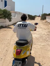 Remera marrón de corte holgado con estampado en la espalda que incluye la palabra "ITTAI" en blanco con líneas rojas y azules, la frase "AN IBIZA PRODUCE" y un texto más pequeño que dice "Ittai Stars Department Gallery - Ibiza Edition. If you're in Ibiza, 112 is not an option.".