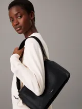 Cartera de mano Calvin Klein con correa ajustable para el hombro. El material exterior es de piel sintética negra con un patrón texturizado de monograma repetido. Presenta una solapa con cierre magnético y un logo metálico plateado en la parte frontal.