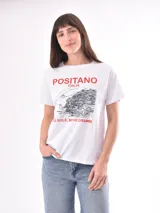 Remera blanca de algodón con cuello redondo y mangas cortas. Presenta un estampado frontal con la inscripción 'POSITANO ITALIA' y 'PIÙ SOLE, MENO DRAMMI' en color rojo, junto a una ilustración lineal en negro de la costa de Positano.