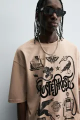 Camiseta de algodón color beige, corte relaxed fit, con cuello redondo y manga corta. Presenta un estampado gráfico grande en negro con motivos variados como una estación de servicio antigua, un caballo alado, una polilla, un pájaro y el texto "RUSTED ROSE".