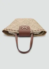 Bolso de mano y hombro de fibra natural trenzada tipo paja, con detalles de cuero sintético marrón, doble asa de hombro, solapa frontal con cierre metálico dorado y forro interior de tela.