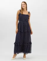 Vestido largo azul marino con estampado de lunares blancos, confeccionado en gasa fluida. Cuenta con tirantes finos ajustables con lazos en los hombros, escote con volados, corte en la cintura con lazo ajustable, y falda larga con múltiples capas y volados horizontales.