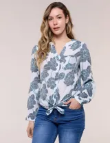 Blusa estampada marca Zac & Rachel, de color blanco con diseño floral en tonos grises y celestes. Posee cuello a la base con escote en V, cierre frontal con botones y un bolsillo aplicado en el pecho. Las mangas son largas con largo regulable mediante presilla y botón, y el ruedo cuenta con terminación anudada al frente.