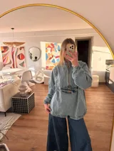 Buzo tipo canguro de denim con diseño oversize. Presenta cuello alto con cierre frontal, bolsillo delantero con cierre, puños elásticos y ruedo regulable con cordón.