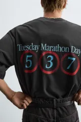 Camiseta negra con efecto lavado, corte boxy fit, cuello redondo y manga corta. Presenta un estampado frontal con texto en tonos turquesa, rojo y blanco que dice "Tuesday 600 Marathon Day Hyde Park".