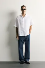 Camisa regular fit confeccionada en tejido de lino, color blanco, con cuello solapa, manga corta y cierre frontal de botonadura.