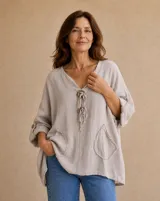 Blusa de lino color beige, de corte holgado y diseño asimétrico más largo en la espalda. Cuenta con escote en V con apliques de arandelas de madera y tiras de tela colgantes, mangas largas arremangables con presilla y botones, y dos bolsillos frontales appliqués con bordes deshilachados.