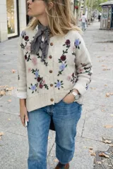 Cardigan de punto color marfil, con cuello redondo a la base, mangas largas y cierre frontal con botones. Presenta un delicado bordado artesanal de flores en tonos bordó, rosado y celeste con tallos y hojas verdes en el frente y las mangas.