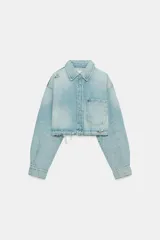 Cazadora corta de denim en tono celeste claro, con cuello solapa, manga larga y cierre frontal con botones. Presenta un bolsillo de plastrón en el delantero y detalles de rotos en el cuello y el ruedo deshilachado.