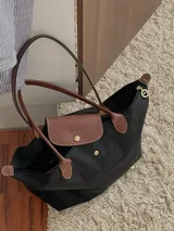 Bolso tipo shopper de cuerpo plegable en color crema o blanco roto, con asas y solapa de cierre en cuero marrón texturizado. Presenta un bolsillo exterior frontal y cierre principal con broche metálico.