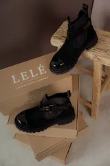 Bota tipo Chelsea de cuero marrón con suela track negra de goma. Presenta paneles elásticos laterales y una costura reforzada en la puntera. Incluye una correa de cuero desmontable con hebilla metálica.