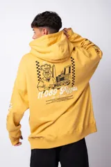 Buzo canguro amarillo ocre oversize de algodón rústico con capucha, cordones negros con letras blancas, bolsillo frontal y puños y cintura acanalados. Presenta estampados en el frente y las mangas.