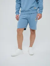 Bermuda de felpa de algodón color celeste, con bolsillos laterales y detalle de costuras verticales al frente. Presenta un parche distintivo de la marca en la pierna izquierda.