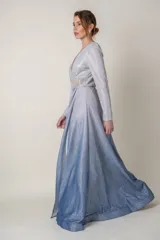 Vestido largo de lurex con acabado brillante en tono lila, con escote en V profundo, mangas largas y cintura marcada con un aplique bordado. La falda presenta un diseño drapeado que nace desde la cintura.