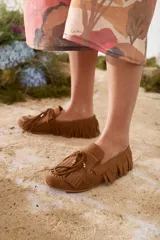 Mocasines de gamuza color tostado, con diseño clásico, flecos alrededor del calzado y en la capellada, y cordón decorativo con detalles metálicos dorados.