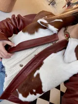 Campera de cuero color marrón rojizo (whisky) con detalles de piel de vaca (marrón y blanco) en el cuello y solapas. Se observa que está abierta, revelando un sweater de punto color crema con botones dorados debajo.