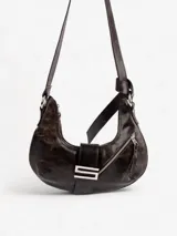 Cartera de cuero genuino color negro, con forma de media luna (hobo bag). Cuenta con asa regulable para llevar al hombro o cruzada, cierre principal de cremallera y broche imán. Presenta un herraje metálico distintivo en forma de "S" en la correa frontal.