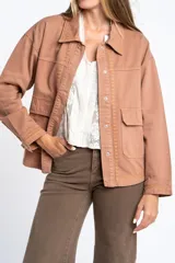 Sobrecamisa de algodón color marrón claro, con cuello camisero y cierre frontal de botones a presión. Presenta detalles de bordado vertical en el frente y en los puños.