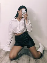 Camisa cropped negra con cuello clásico, mangas largas con puños abotonados y cierre frontal con botones. Presenta detalle de tachas plateadas en el cuello.