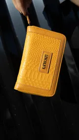Billetera mediana con textura tipo croco, color mostaza, con cierre perimetral y logo de la marca grabado en el frente.