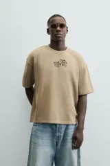 Sudadera de manga corta color beige, con cuello redondo y corte relajado. Presenta un bordado y estampado en el pecho con texto y un diseño floral.