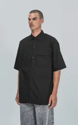 Camisa de manga corta en popelina de algodón, con diseño oversize y cierre frontal de botones. Presenta dos bolsillos delanteros, uno con solapa y otro con ojal y botón.