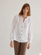 Camisa de corte recto y manga larga, confeccionada en algodón blanco. Presenta un canesú superior con bordado inglés floral y detalle de placa metálica con logo en la espalda.