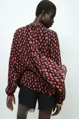 Camisa de cuello subido y manga larga con puños elásticos. Presenta un diseño de lunares en tonos fucsia y amarillo sobre fondo negro, con detalle de pliegues en el frente y cierre de cremallera oculta en la espalda.