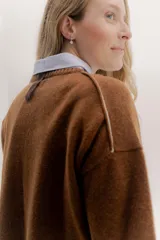 Buzo de lambswool color beige, con escote redondo y detalle en V. Presenta cierres metálicos en los puños que revelan un interior en contraste de color marrón y ribete rojo. Tiene tajos laterales para mayor comodidad y un fit amplio.