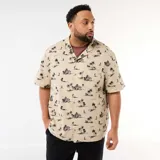 Camisa de manga corta color beige con estampado tropical de palmeras y veleros en tonos oscuros, cuello cubano y un bolsillo aplicado en el pecho.