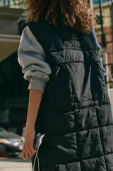 Chaleco largo acolchado de color negro, con cierre frontal completo y diseño sin mangas. Presenta un acabado matelasse y un borde contrastante en color claro a lo largo de la cremallera.