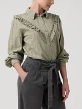 Blusa verde de algodón orgánico con cuello camisero, manga larga y cierre frontal de botones. Presenta volantes bordados que recorren el frente, la espalda y las sisas, además de detalles bordados en toda la prenda.