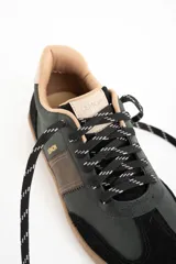 Zapatilla urbana de caña baja, con capellada combinada en tonos verde oscuro, marrón y negro. Presenta puntera y talón en gamuza negra, detalles en cuero marrón en el lateral y forro interior color beige. La suela es de goma color caramelo.