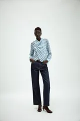 Camisa de manga larga con cuello fluido y diseño cruzado en el frente. Confeccionada en tejido semitransparente de color celeste con estampado de lunares negros. Presenta bajo asimétrico y cierre en la espalda con botones forrados.