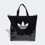 Bolso shopper Adidas de diseño minimalista en material sintético efecto charol brillante color negro. Cuenta con doble asa de mano y correas largas para llevar al hombro, cierre principal y logo del trifolio estampado en color blanco en la parte frontal.