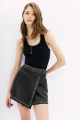 Falda corta negra de cuero ovino con diseño cruzado tipo wrap. Presenta apliques de tachas y piedras plateadas y doradas a lo largo de los bordes y en la cintura.