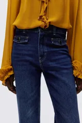 Jean de tiro alto con corte acampanado desde la rodilla. Presenta cierre frontal con doble botón, bolsillos frontales decorativos con solapa y botón, y bolsillos traseros tipo parche.