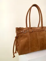 Bolso grande de cuero marrón con textura tipo cocodrilo, estructura rígida y asas para llevar al hombro. Tiene una tira con tachas y hebillas en cada extremo, también lleva lazos en los costados.